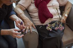 Vrouw lakt nagels van een oudere vrouw in een stoel met paarse nagellak. De oudere vrouw heeft bloemen in haar hand.