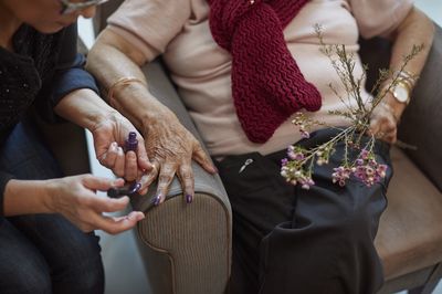 Vrouw lakt nagels van een oudere vrouw in een stoel met paarse nagellak. De oudere vrouw heeft bloemen in haar hand.