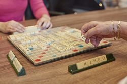 Een scrabblebord met gelegde woorden erop en drie donkergroene plankjes met letters. Een gerimpelde hand met twee gouden schakelarmbanden legt de letter 'G' op het bord