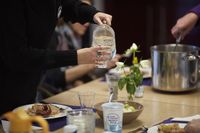 Een gedekte tafel met pannen en borden met eten. Iemand schenkt met een waterkan een glas water in.