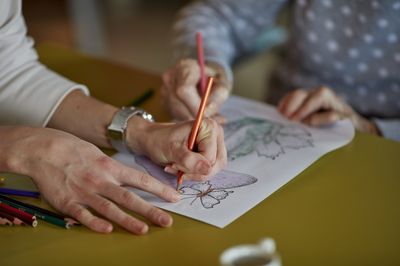 Vier handen houden een kleurplaat vast met paddenstoelen en vlinders. Twee van de handen hebben een oranje en roze kleurpotlood vast en kleuren de kleurplaat in