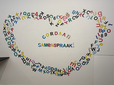 Allemaal gekleurde letterstickers op de muur geplakt in de vorm van een hart. In het midden staat de tekst geplakt Cordaan Samenspraak!