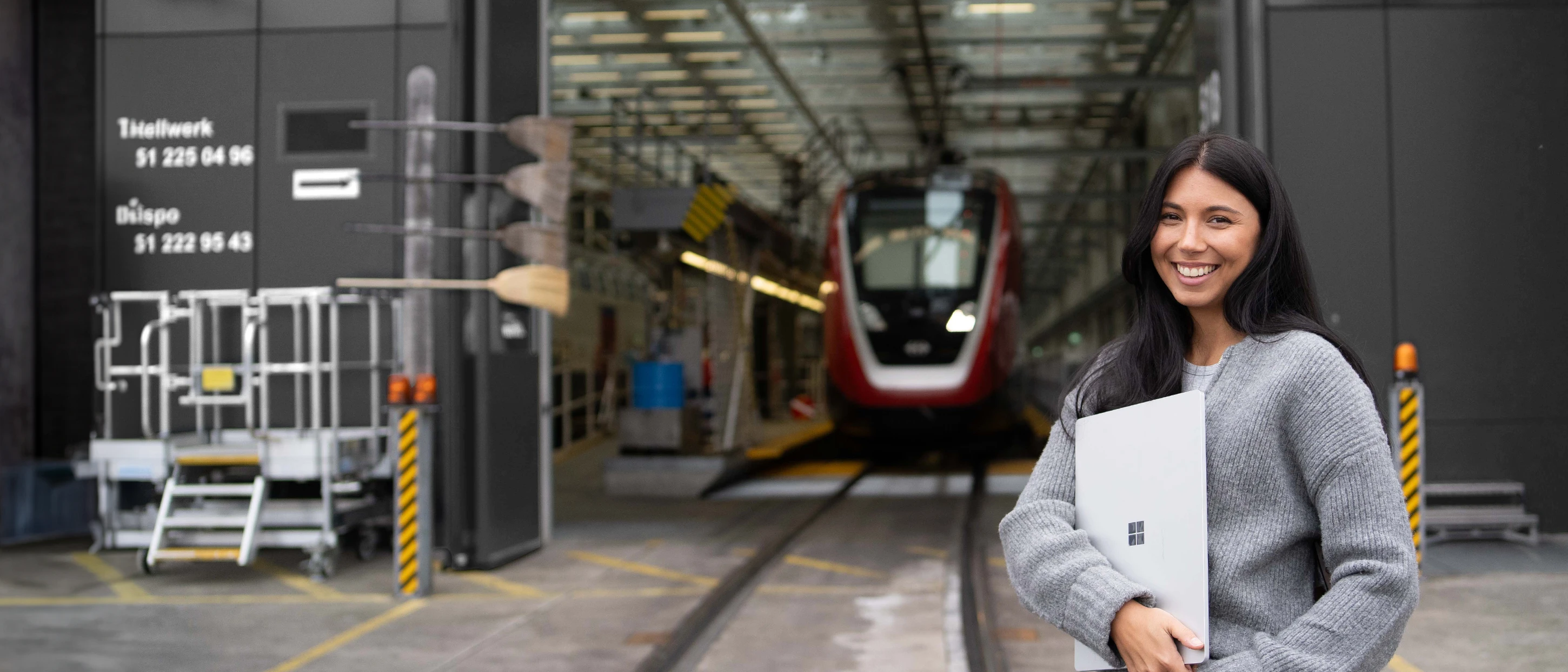 Eine junge Frau mit Laptop lächelt vor einem modernen Zug in einem Bahnbetriebswerk.