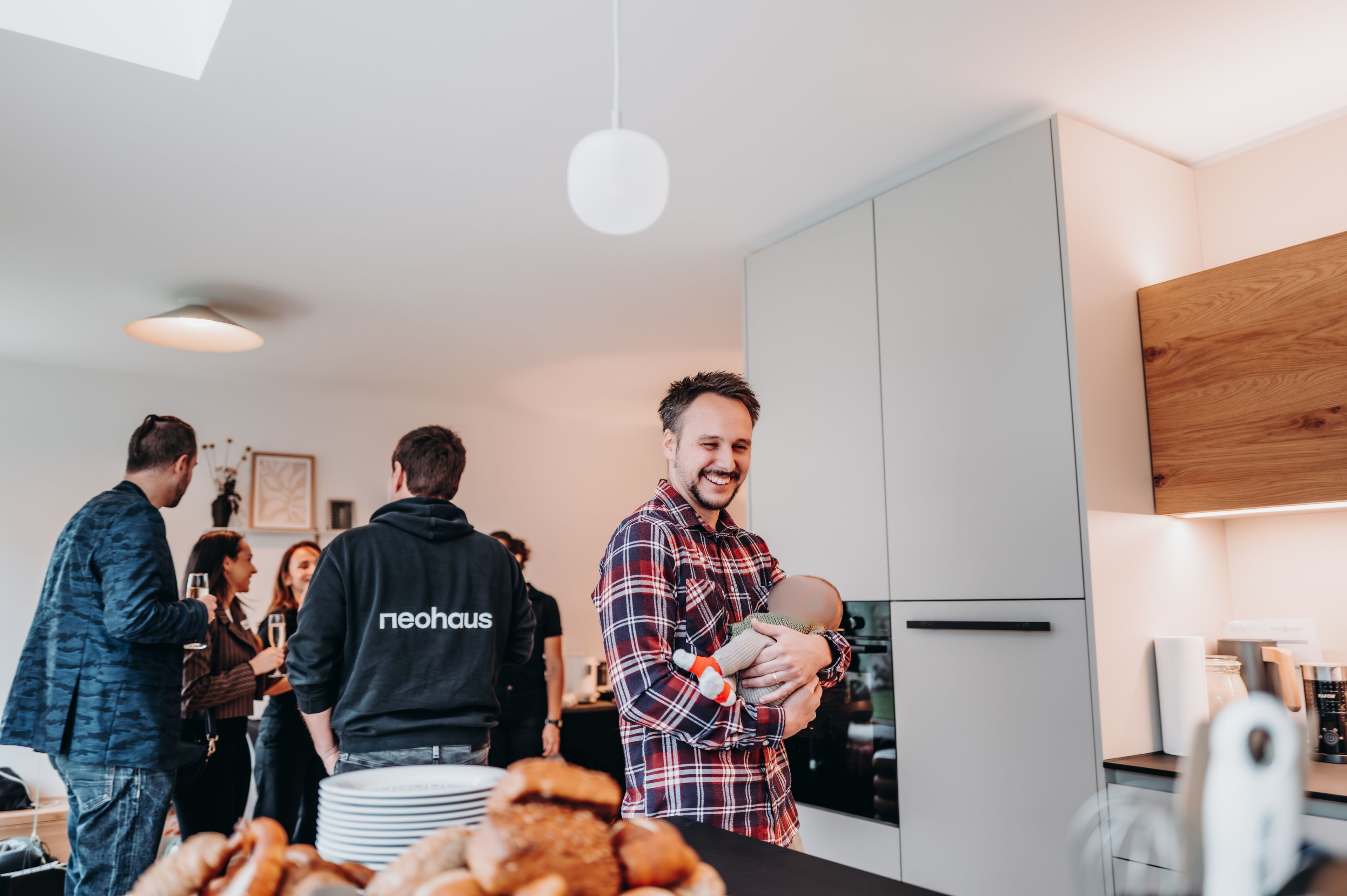 A smiling man holding a baby stands in a kitchen with a group of people chatting in the background. Plates of pastries are on the counter.