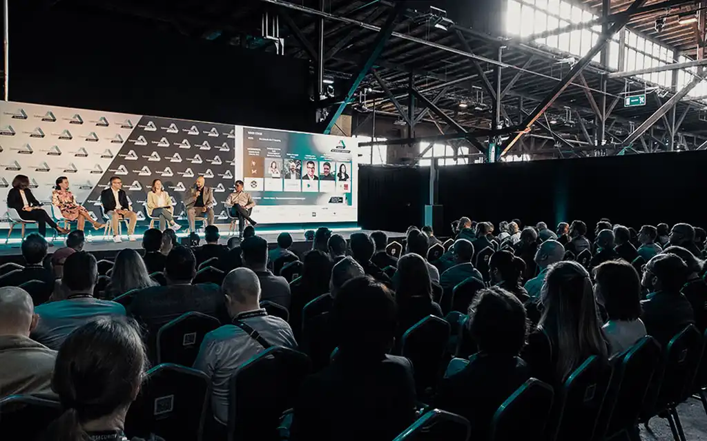 Menschen auf Stühlen vor einer Vortragsbühne mit vortragenden Personen