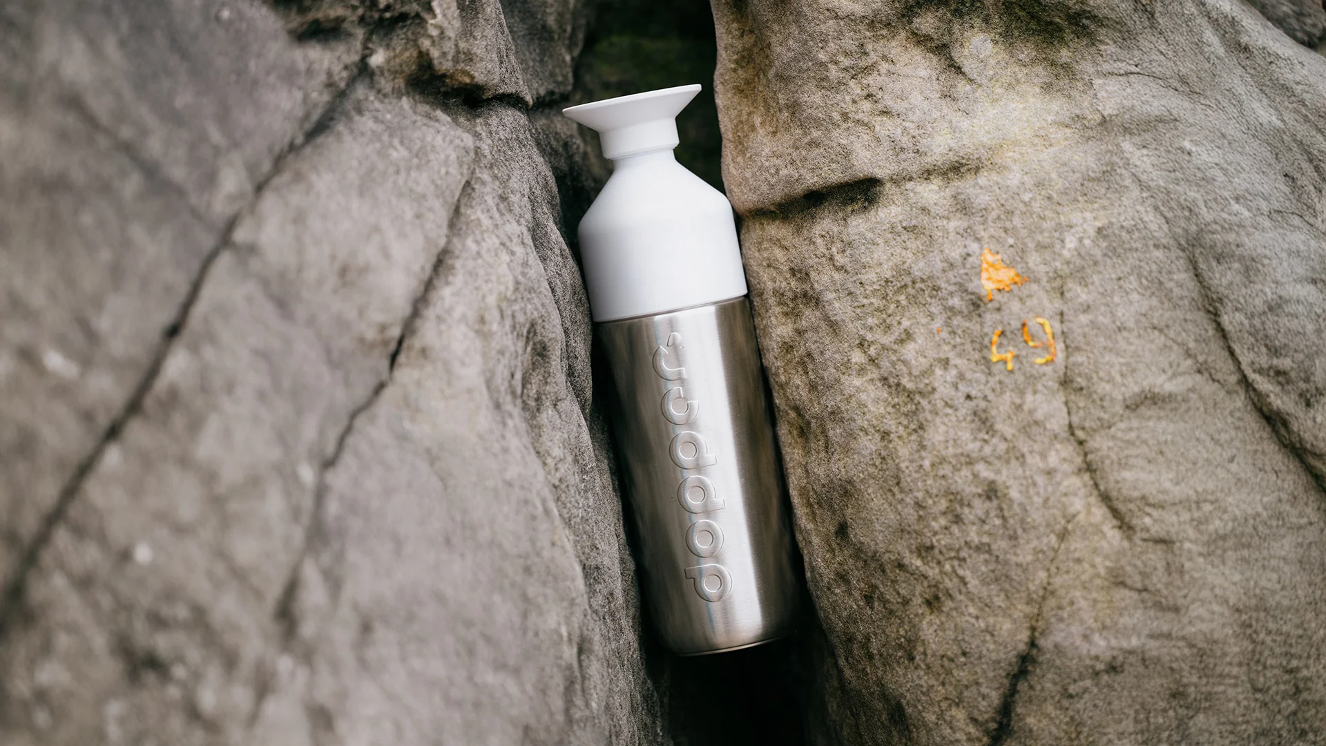 An Insulated Silver Dopper water bottle with a white cap is wedged between two large gray rocks.