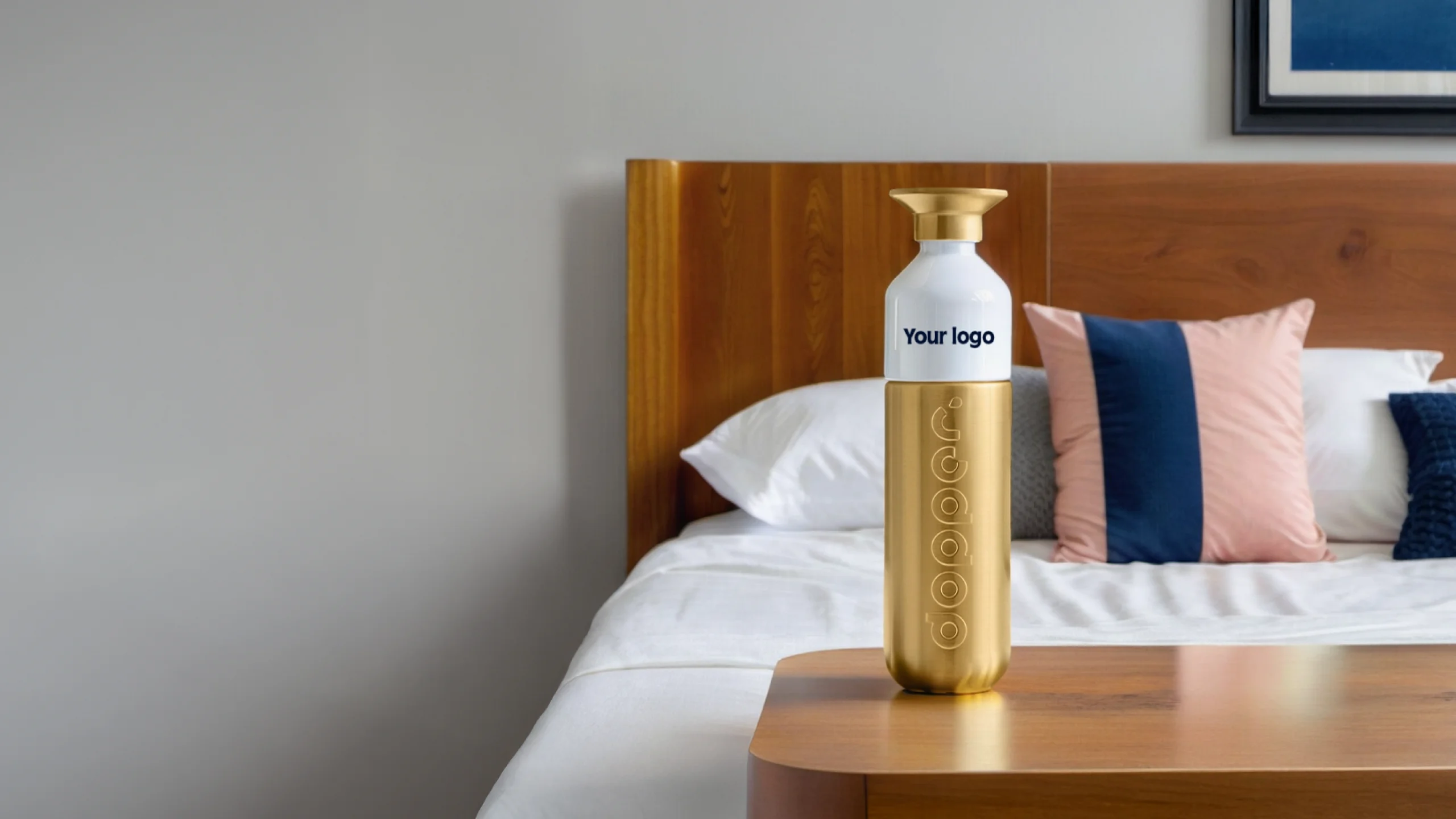 A gold and white bottle with "Your logo" text on a wooden bedside table, next to a neatly made bed with decorative pillows.