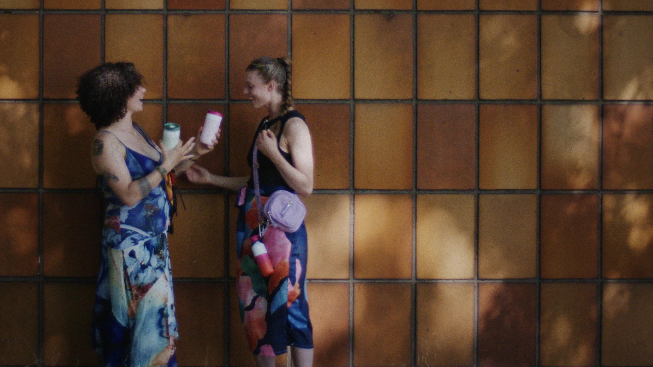 Two people in colorful outfits laugh and share a drink in a Dopper Travel Mug in front of a sunlit tiled wall.