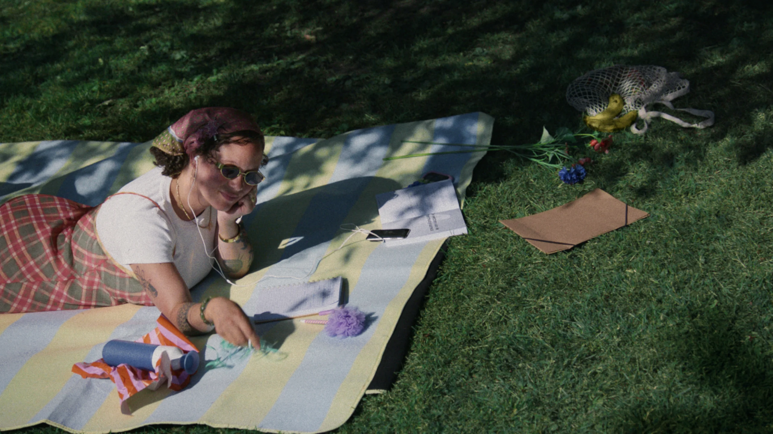 A woman lies on a striped blanket reading under a tree. She wears sunglasses and a headscarf, with flowers and papers beside her on the grass, and a blue Dopper Original bottle.