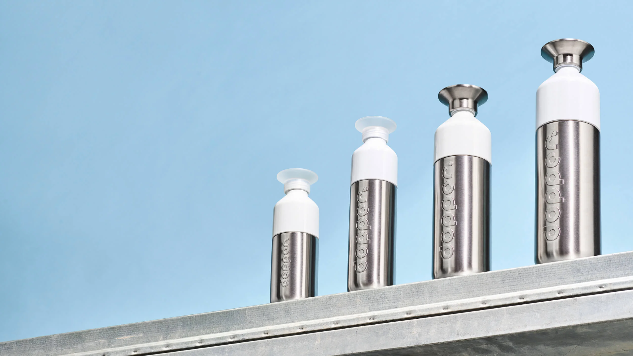 Four stainless Dopper steel water bottles with white tops arranged in ascending order on a metal surface against a blue background.