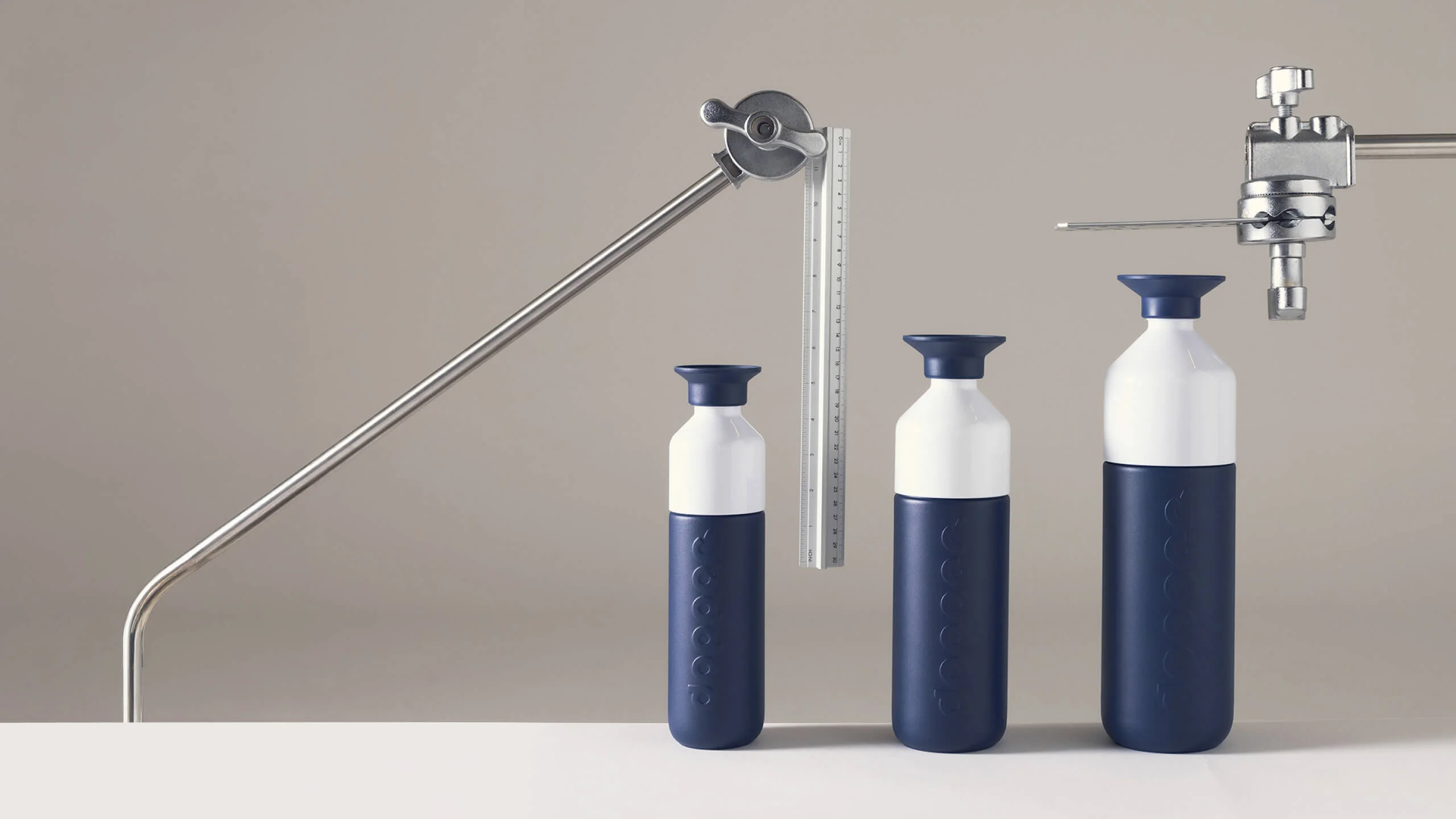 Three navy-blue Dopper water bottles in ascending sizes, with white bodies and navy caps, displayed alongside a ruler and industrial measuring equipment on a neutral background