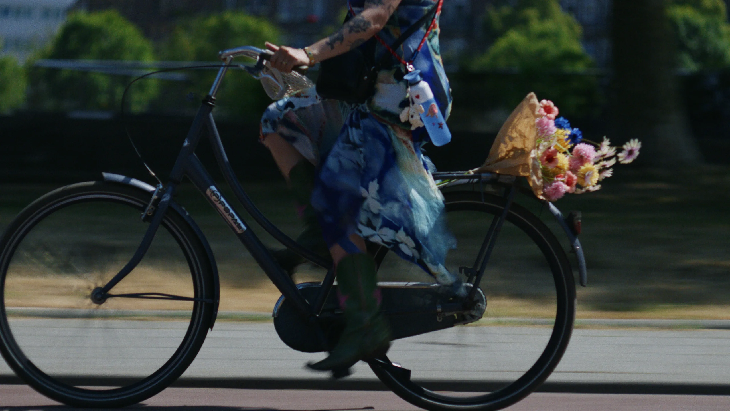 Person cycling outdoors with a bouquet of flowers attached to the back of the bicycle, wearing colorful attire and green boots and a red Dopper Cord holding a blue Dopper Original bottle with stickers.