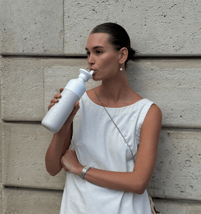 A woman sips from from a Dopper Insulated Gentle Grey water bottle with a Flip Straw, while 3 sentences appear: Hot for 9h Cord for 24h / Leakproof / 90% recycled steel