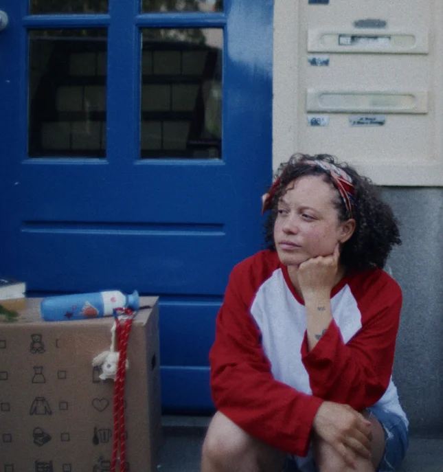 Person in red and white shirt sitting on a doorstep next to a box and a blue Dopper Original water bottle, looking contemplative.