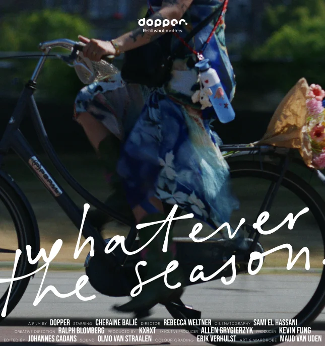 A person riding a bicycle wearing a blue outfit with floral patterns, carrying a blue Dopper Original water bottle, with the text "whatever the season" overlaid.