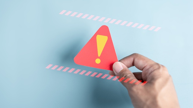 Hand hält ein rotes Warnsymbol in Form eines Dreiecks mit gelbem Ausrufezeichen auf blauem Hintergrund. Für Krisensituationen und Alarm-Kommunikation ist SMS der ideale Kanal.