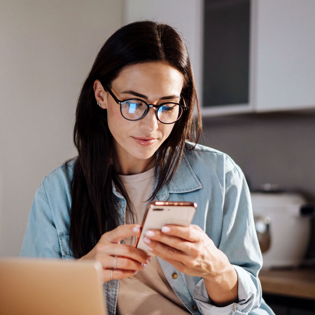 websms - junge Frau zuhause vor dem Laptop mit ihrem Smartphone. Unternehmen können SMS-Nachrichten an Ihre Kunden mit unterschiedlichen Absendernummern und Namen versenden.