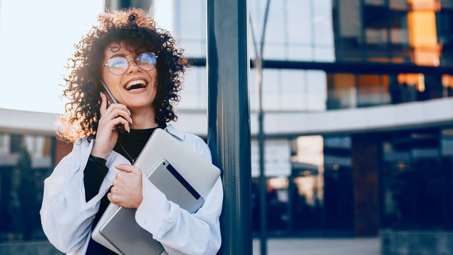 Person steht im Freien vor einem modernen Bürogebäude und telefoniert. Dank der KI-Unterstützung von aiTel sind Unternehmen rund um die Uhr erreichbar und Standardfragen werden automatisch beantwortet.