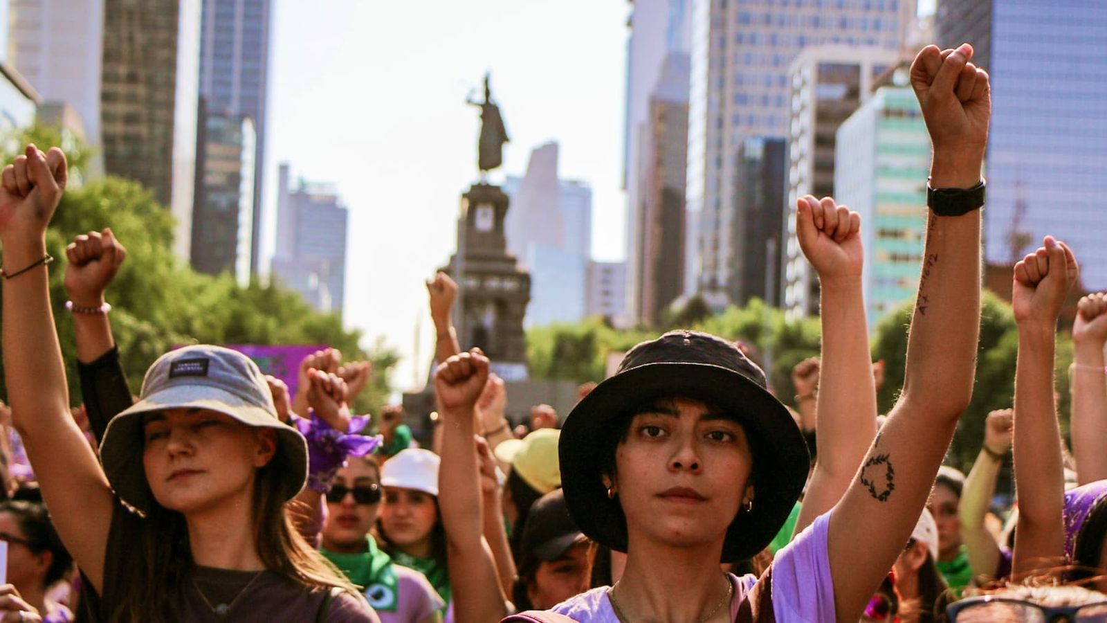 A crowd of women standing in a street with their arms raised in the air and fists clenched, in front of a blurred statue and surrounded by high-rise buildings.