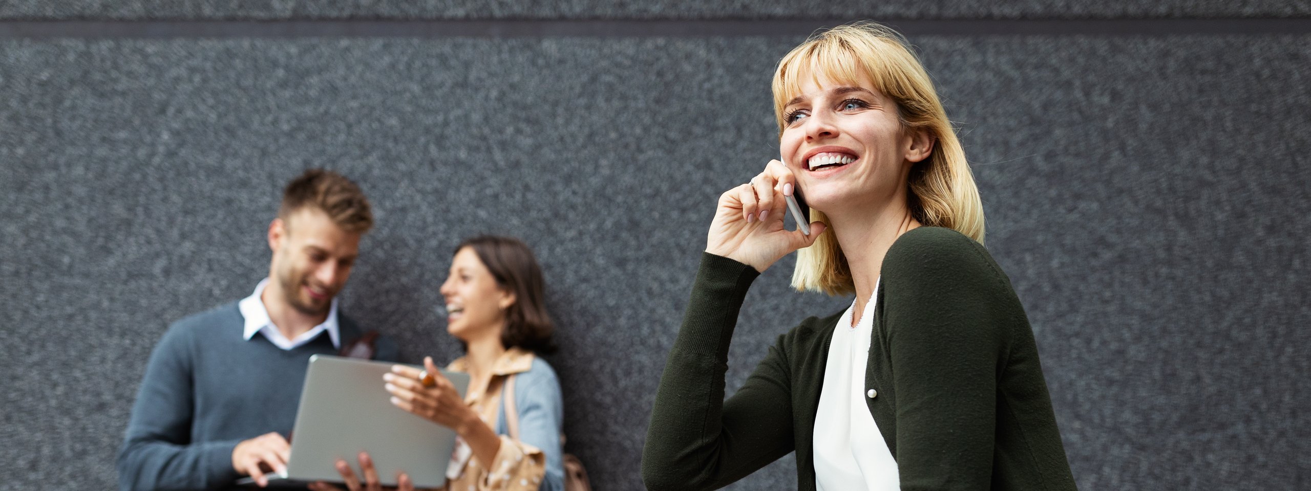 Woman on a bike talking on the phone, smiling, while a man and woman in the background look at a laptop and laugh.
