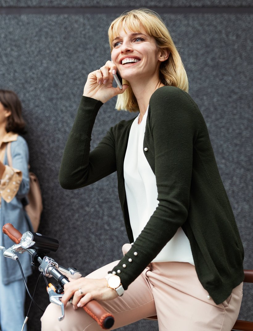 Woman on a bike talking on the phone, smiling, while a man and woman in the background look at a laptop and laugh.