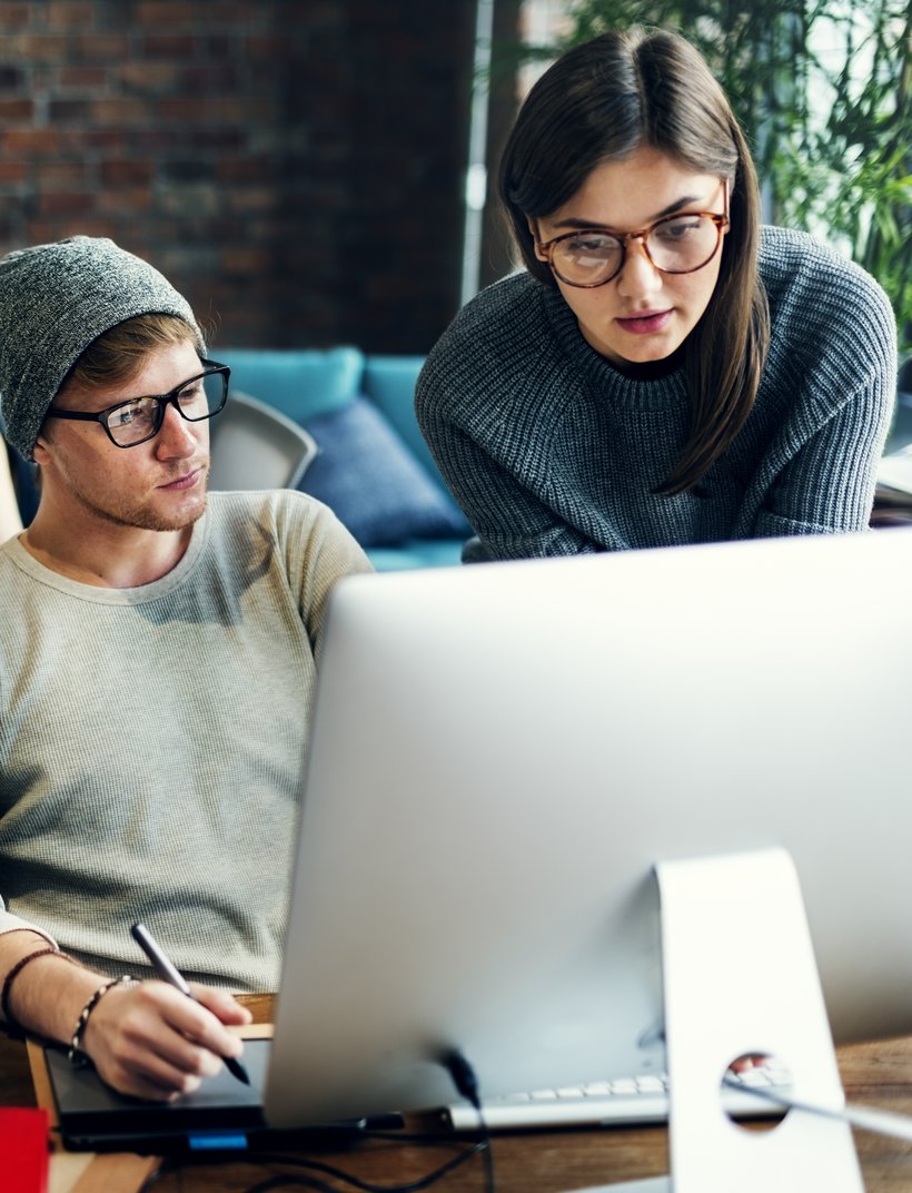 Zwei Personen arbeiten gemeinsam an einem Computer in einem modernen Büro, im Hintergrund sitzt eine weitere Person an einem Tisch mit Laptop.