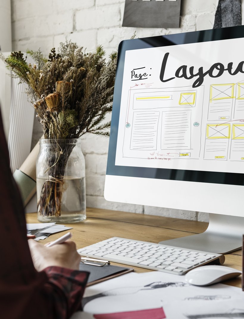 Person arbeitet am Computer an einem Webseiten-Layout-Entwurf auf einem Schreibtisch mit Tastatur und Notizen.