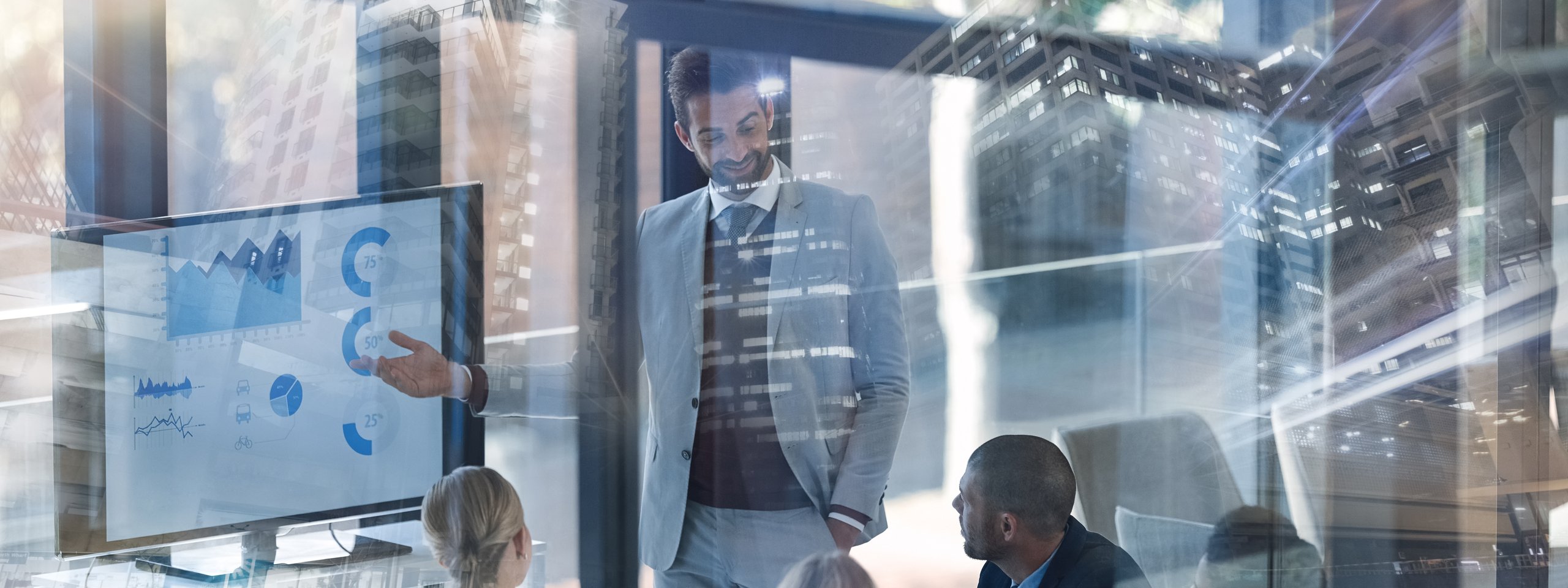 Business meeting in a glass-walled conference room with a man presenting data on a screen, reflections of city buildings visible.