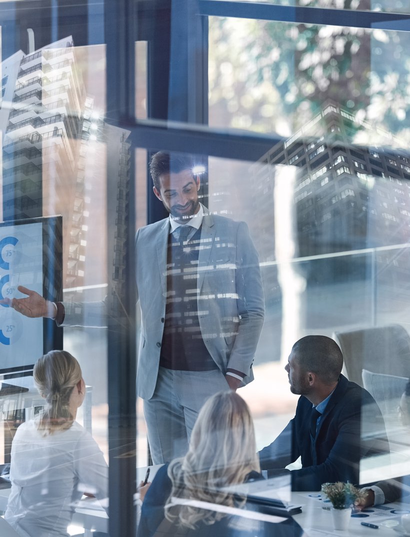 Business meeting in a glass-walled conference room with a man presenting data on a screen, reflections of city buildings visible.