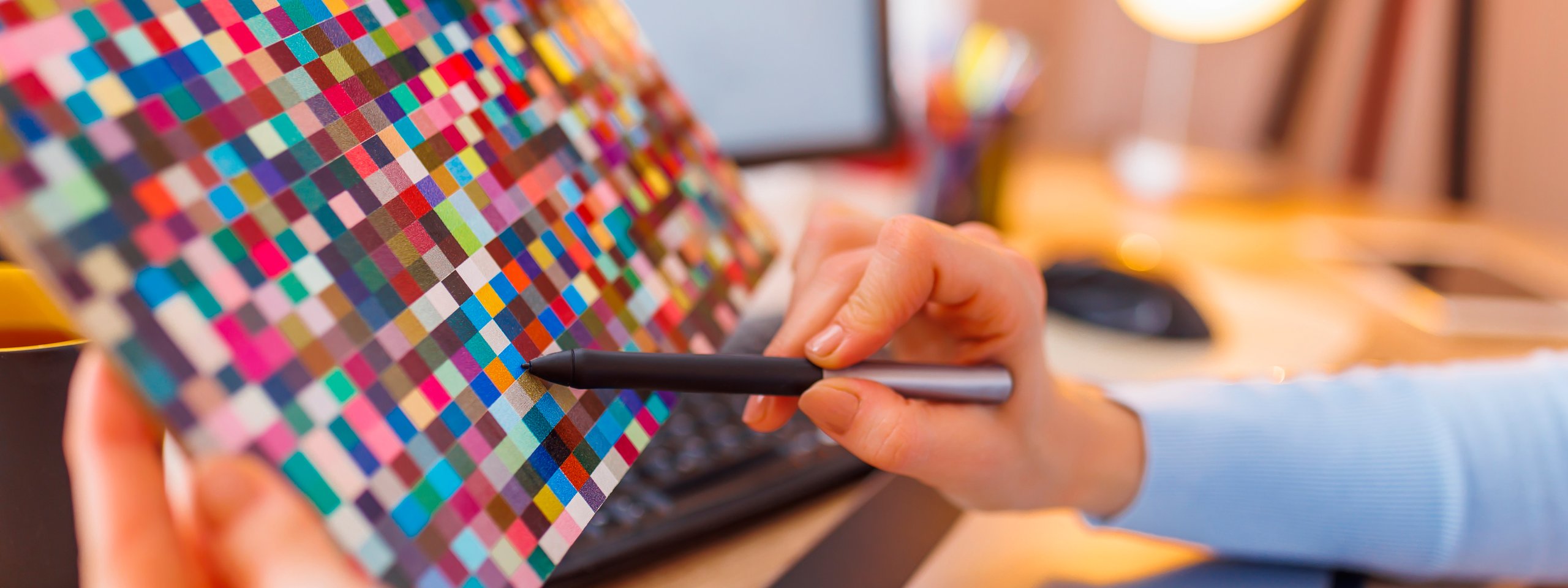 Designerhand hält eine Farbpalette aus bunten Pixelquadraten und zeigt mit einem Stift darauf, vor einem Computerarbeitsplatz.