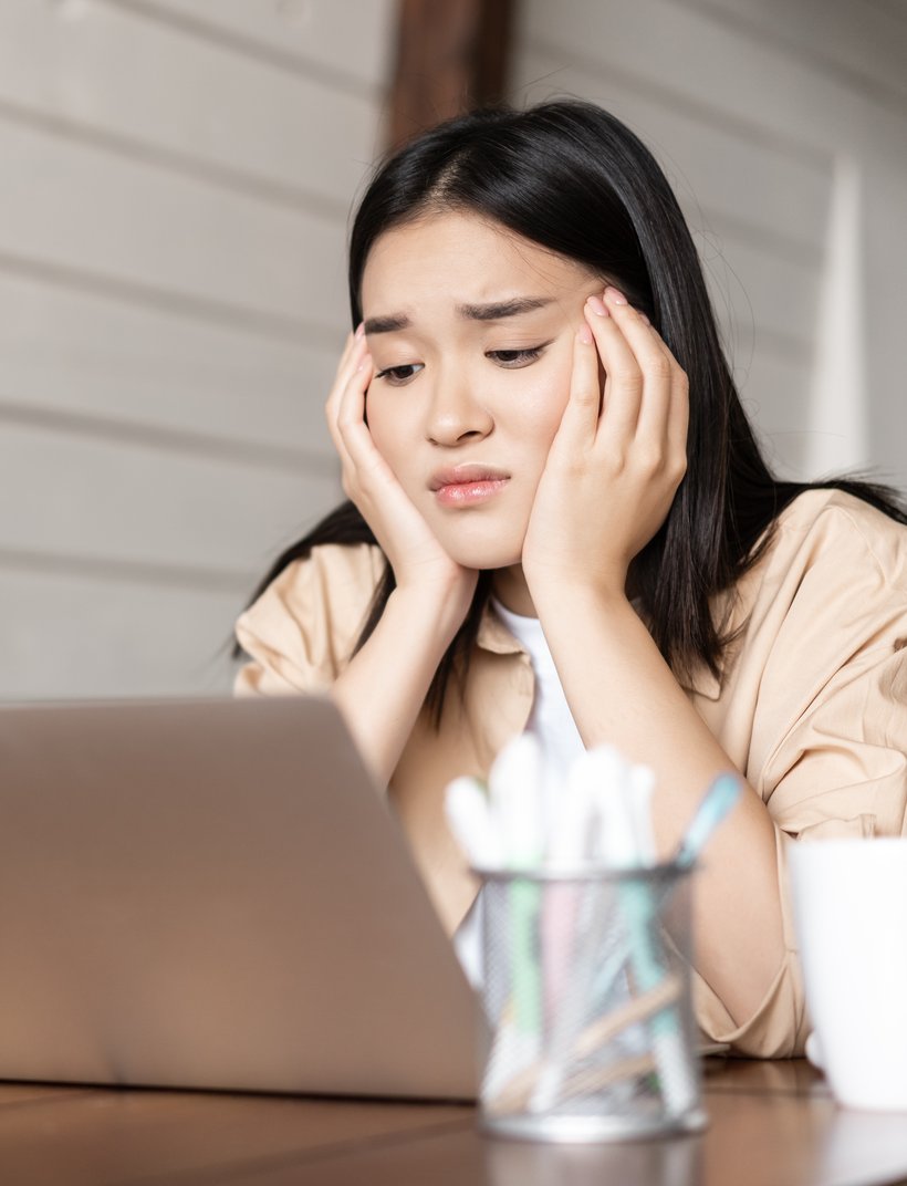 Junge Frau sitzt erschöpft vor einem Laptop und stützt nachdenklich den Kopf mit beiden Händen.