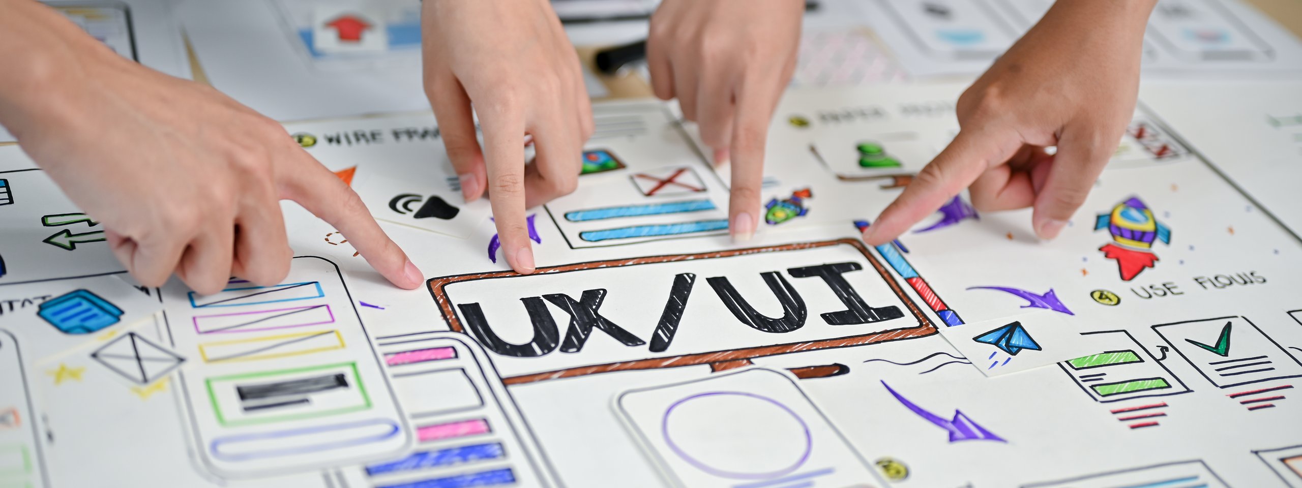 Mehrere Hände zeigen auf skizzierte UX/UI-Wireframes mit dem Schriftzug „UX/UI“ auf Papier.