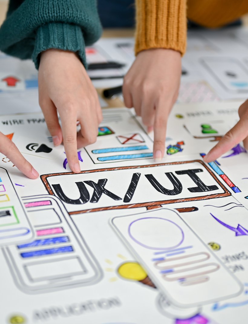Mehrere Hände zeigen auf skizzierte UX/UI-Wireframes mit dem Schriftzug „UX/UI“ auf Papier.
