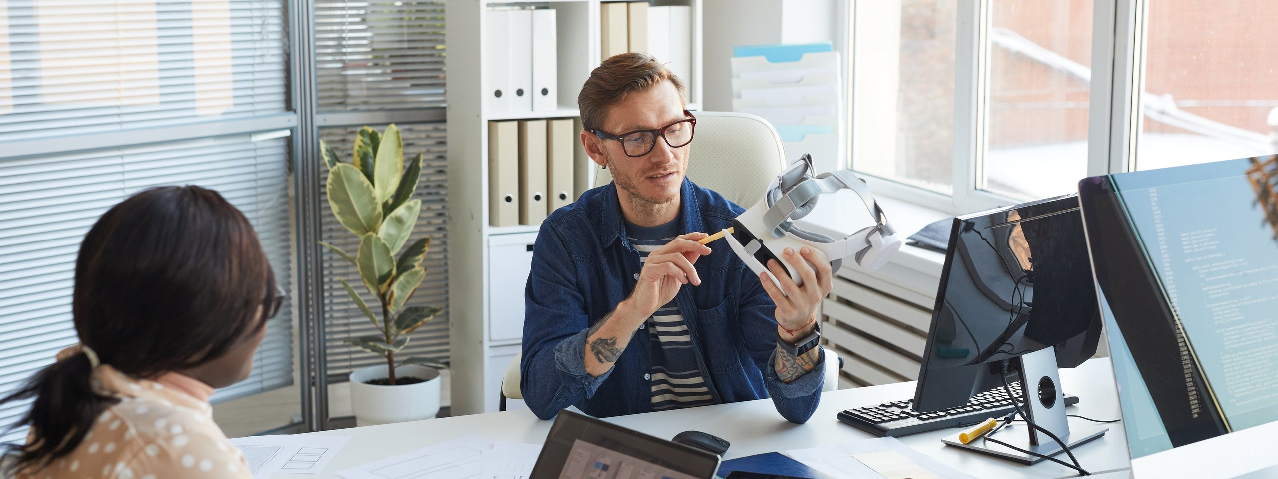 Zwei UX-Designer im Büro besprechen ein VR-Headset, umgeben von Laptops, Monitoren und Skizzen auf dem Schreibtisch.
