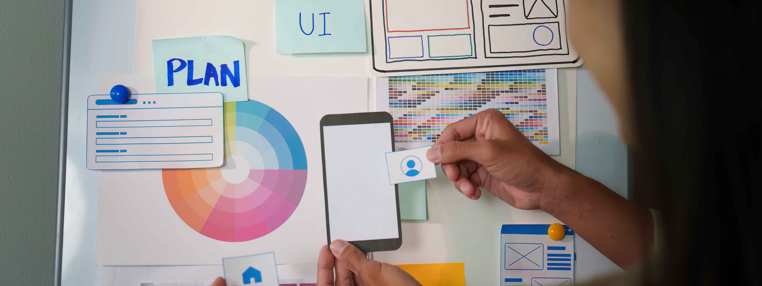 Person planning UX/UI design with color palettes, wireframes, and Post-it notes on a board.