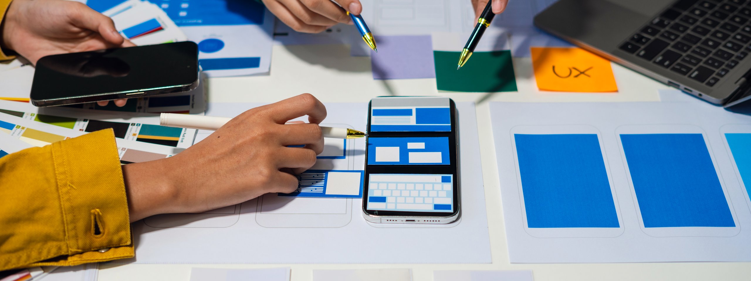 Team entwirft UX-Design: Hände mit Stiften, Smartphone-Mockups, Farbproben und Laptop auf Tisch.