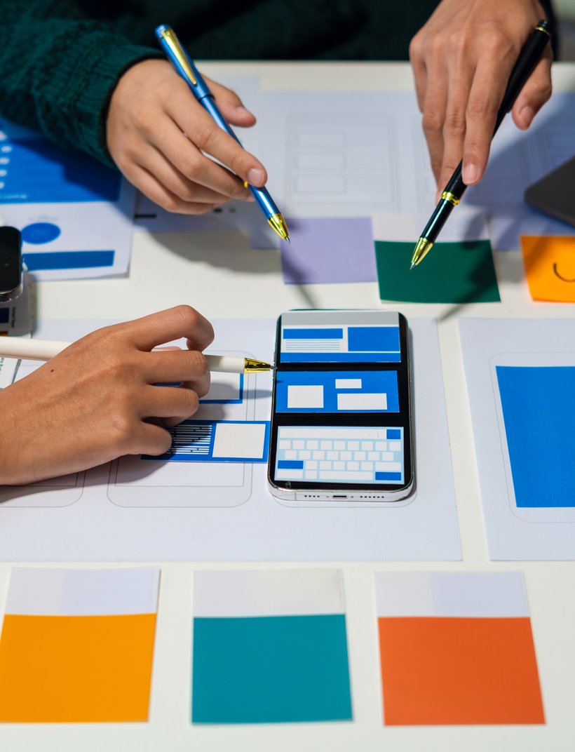 Team entwirft UX-Design: Hände mit Stiften, Smartphone-Mockups, Farbproben und Laptop auf Tisch.