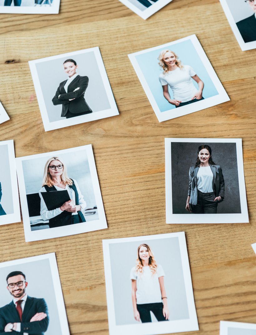 Mehrere Polaroidfotos von verschiedenen beruflich gekleideten Personen liegen auf einem Holztisch verstreut.