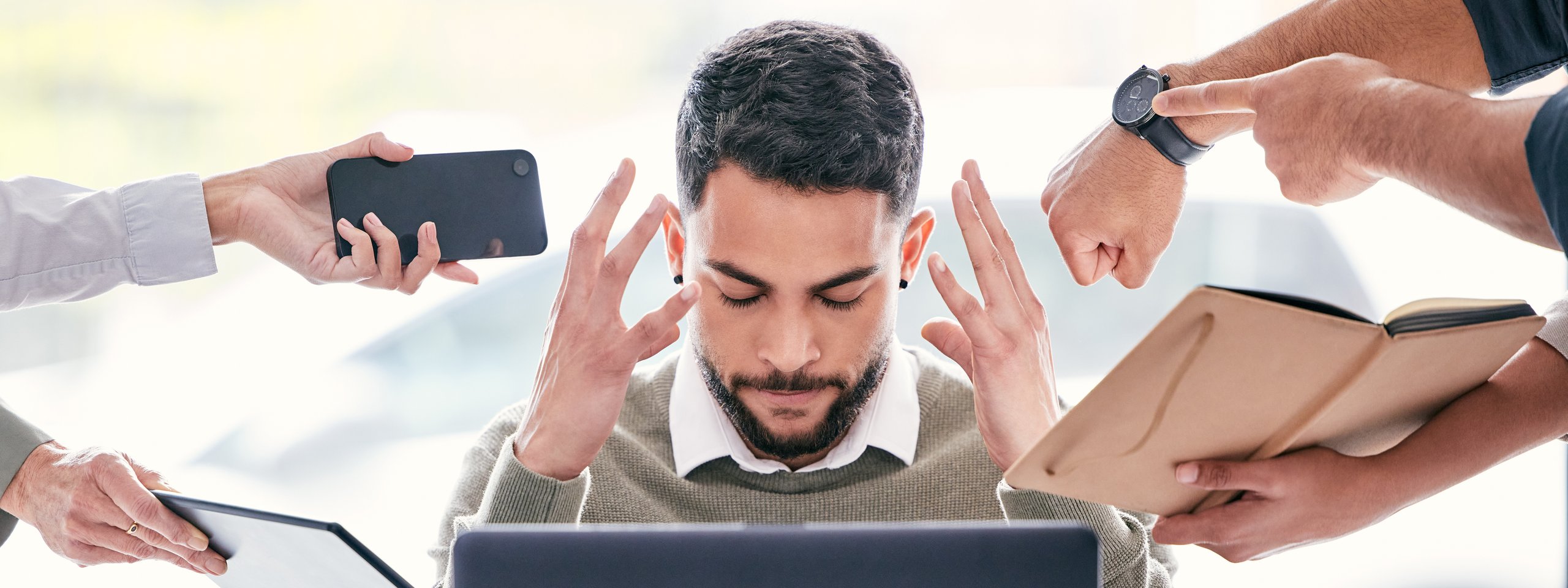 Gestresster Büroangestellter am Laptop, umgeben von fordernden Händen mit Handy, Uhr, Dokumenten und Notizen.