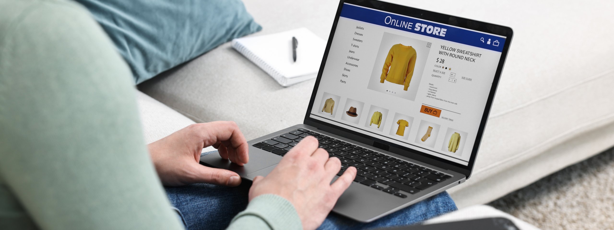 Person shopping online for a yellow sweatshirt on a laptop while sitting on a couch. Nearby, a phone and notebook are visible.