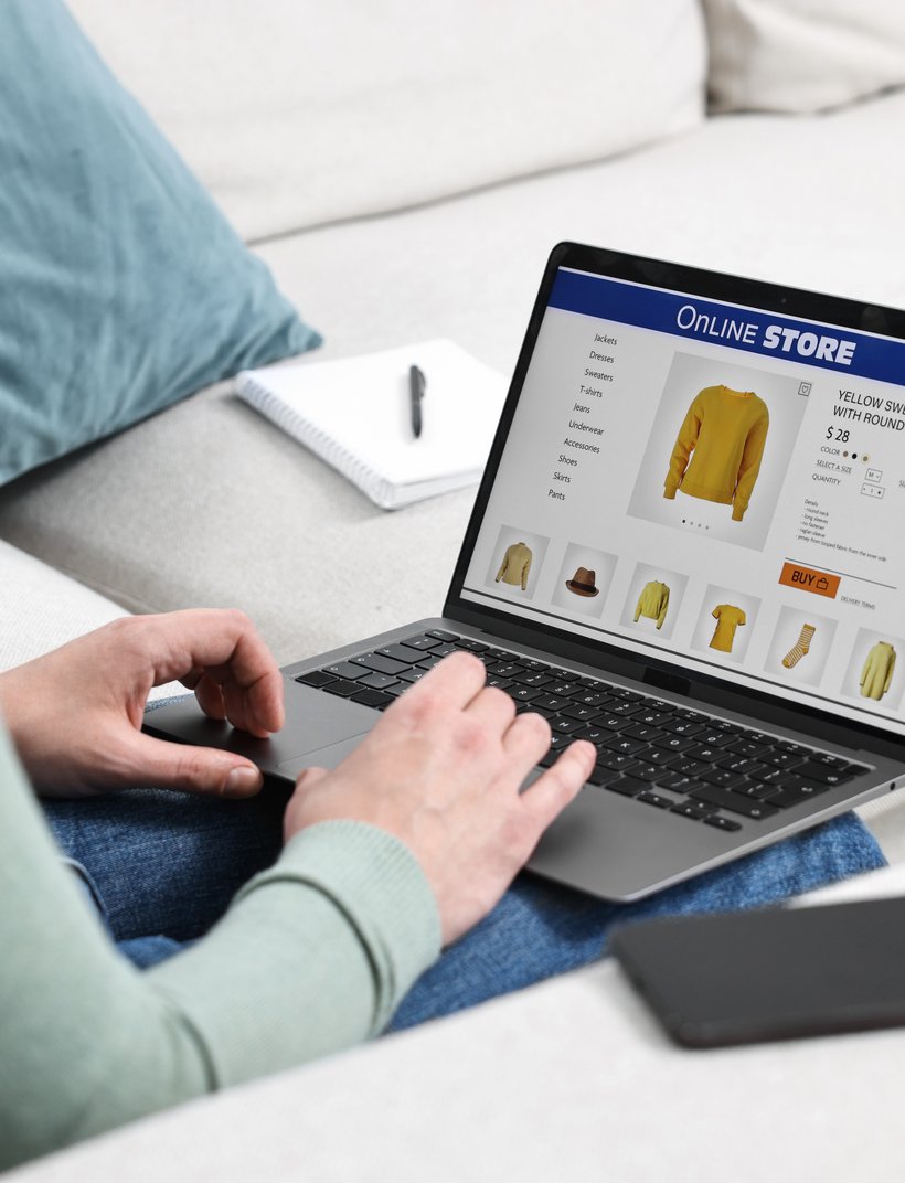 Person shopping online for a yellow sweatshirt on a laptop while sitting on a couch. Nearby, a phone and notebook are visible.