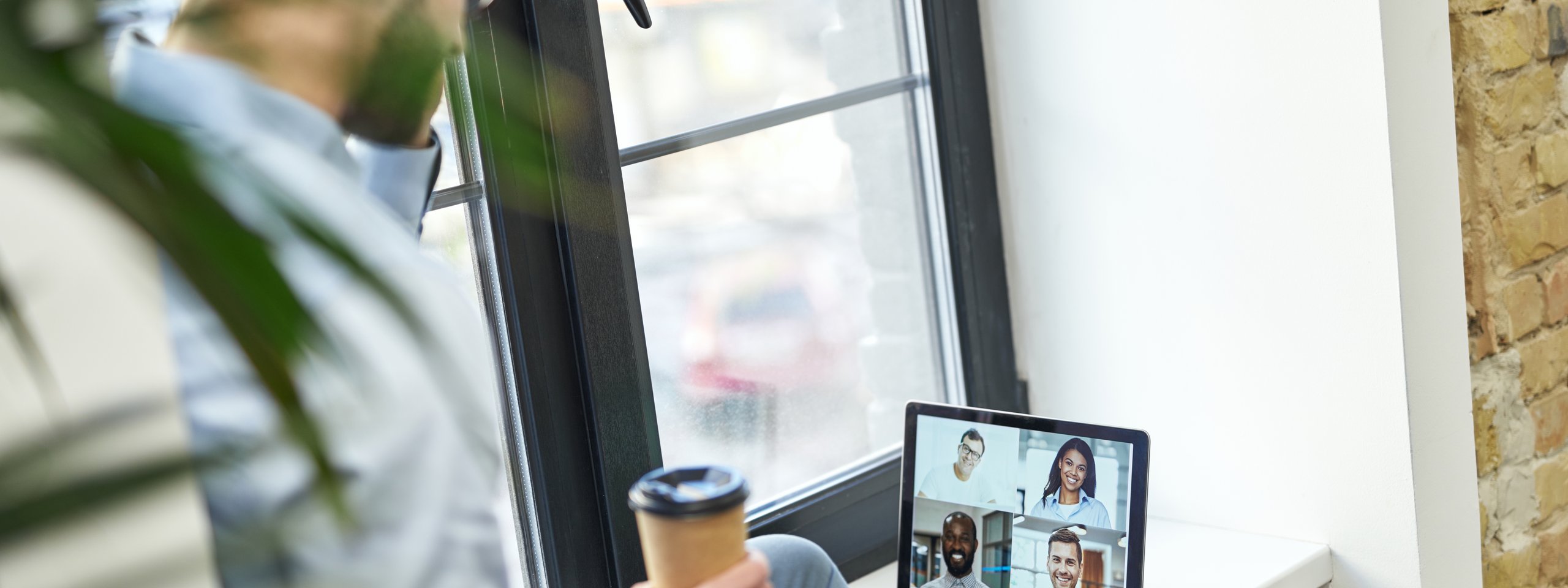 Ein Mann hält einen Kaffeebecher und nimmt an einem Videocall mit vier Kollegen auf einem Laptop am Fenster teil.