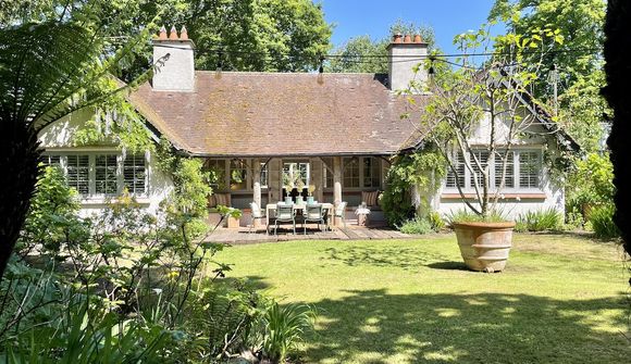 Charming cottage with a sloped roof, surrounded by lush greenery and trees, featuring a patio with outdoor dining furniture.