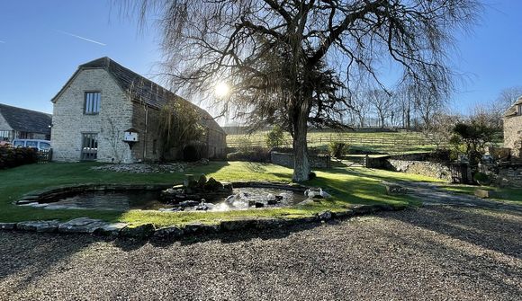 Stone building beside a pond and bare tree on a sunny day, with distant hills and clear blue sky in the background.