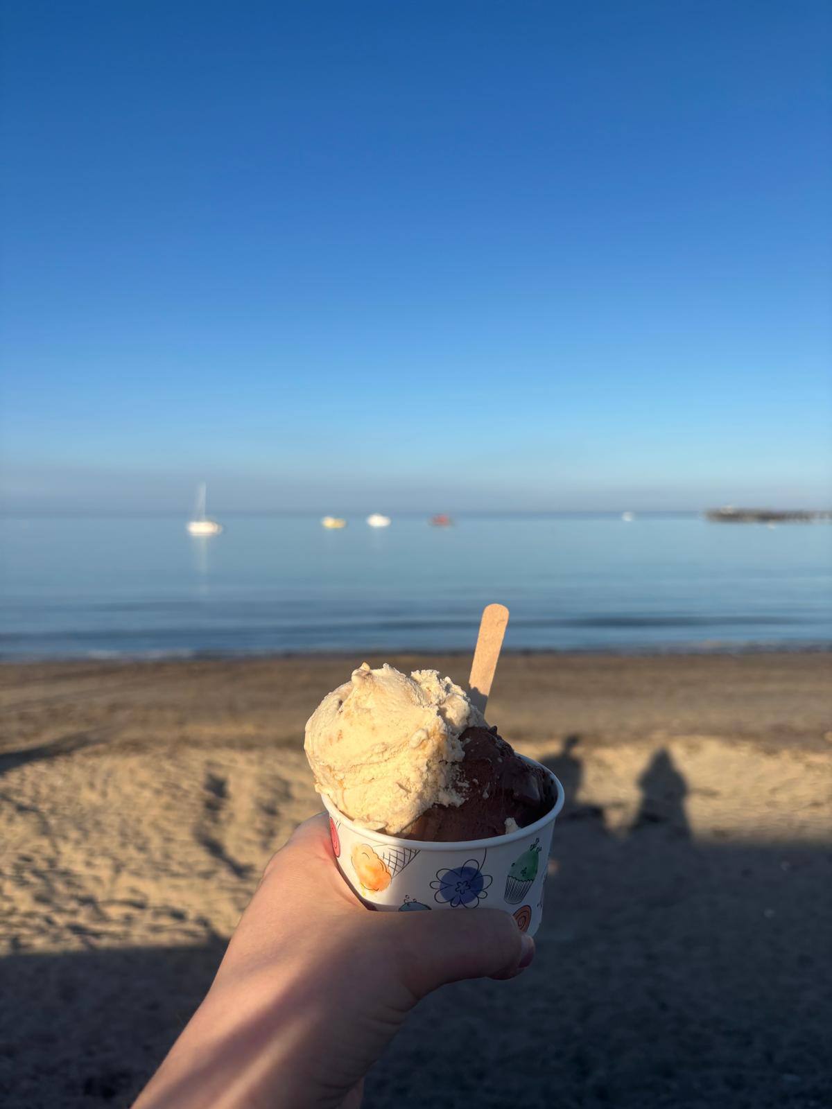 Hand holding ice cream cup with two scoops, against a backdrop of a sandy beach and calm sea under a clear blue sky.