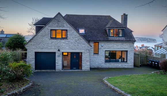 A two-story stone house with a dark roof, large windows, and a garage, set against a backdrop of a sunset and distant water view.