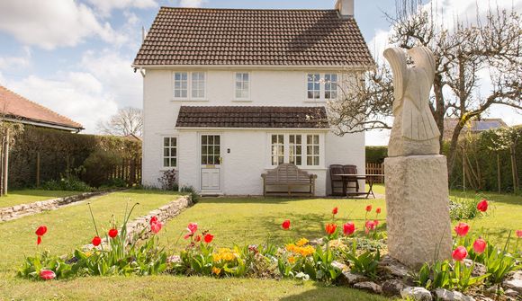 A charming white cottage with a red-tiled roof, surrounded by a garden with red tulips and a stone statue, under a partly cloudy sky.