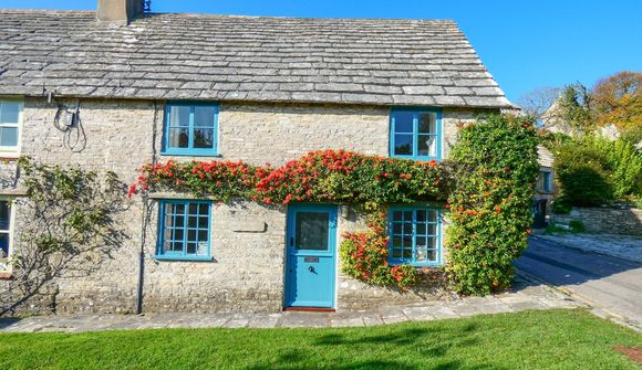 Stone cottage with blue windows and door, adorned with red and green climbing plants, set against a clear blue sky and lush green lawn.