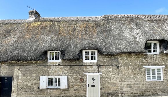 Stone cottage with a thatched roof, white-framed windows, and a light gray door, set against a clear blue sky.