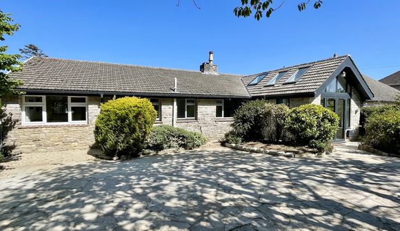 Single-story stone house with a sloped roof, surrounded by shrubs, under a clear blue sky. Shadows of tree branches on the ground.